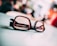 A pair of glasses with black frames resting on a light-colored surface, with text visible on the left lens. The background is blurred, adding depth and focus to the glasses.