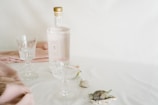 A still life arrangement featuring an elegant bottle with a light pink label and gold cap, alongside two crystal glasses. The setting includes soft pink fabric draped on a white surface, with decorative sea shells adding a touch of nature.