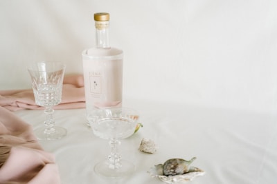 A still life arrangement featuring an elegant bottle with a light pink label and gold cap, alongside two crystal glasses. The setting includes soft pink fabric draped on a white surface, with decorative sea shells adding a touch of nature.