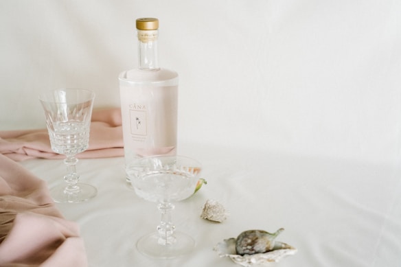 A still life arrangement featuring an elegant bottle with a light pink label and gold cap, alongside two crystal glasses. The setting includes soft pink fabric draped on a white surface, with decorative sea shells adding a touch of nature.