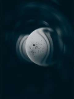 A circular, moon-like object with scattered bubbles of various sizes appears in the center. Surrounding it are swirling shadows and dark textures, creating a mysterious and cosmic effect.