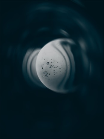 A circular, moon-like object with scattered bubbles of various sizes appears in the center. Surrounding it are swirling shadows and dark textures, creating a mysterious and cosmic effect.
