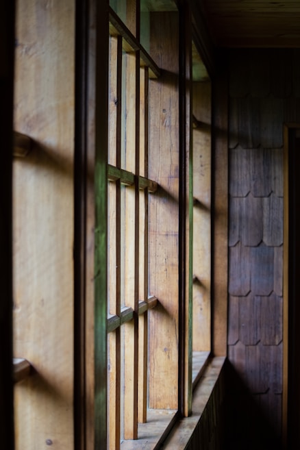 A series of wooden window panels creates an intricate play of light and shadow as the sunlight streams in. The predominantly wooden texture gives a rustic and natural feel, with the window frames casting geometric shadows on the walls.