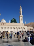 Large gathering of people in front of a mosque with a prominent green dome and a tall minaret, set against a clear blue sky. The crowd appears to be engaged in prayer or reflection, with some seated on the ground and others standing.