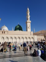 Large gathering of people in front of a mosque with a prominent green dome and a tall minaret, set against a clear blue sky. The crowd appears to be engaged in prayer or reflection, with some seated on the ground and others standing.