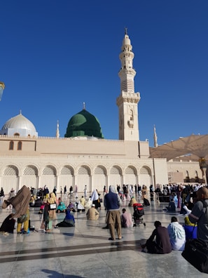 Large gathering of people in front of a mosque with a prominent green dome and a tall minaret, set against a clear blue sky. The crowd appears to be engaged in prayer or reflection, with some seated on the ground and others standing.