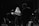 A black and white image of musicians performing on stage. A woman with long hair stands at the microphone, while a man plays a guitar beside her. Another performer sings into a microphone in the background. Musical equipment and stands are visible around them.