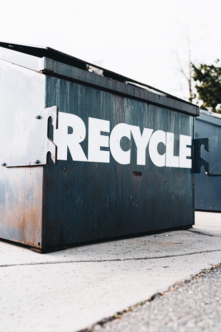 A large metal recycling container with the word 'RECYCLE' prominently displayed in white capital letters. The container shows signs of rust and wear, suggesting regular use. It is situated on a concrete surface, with trees visible in the background.