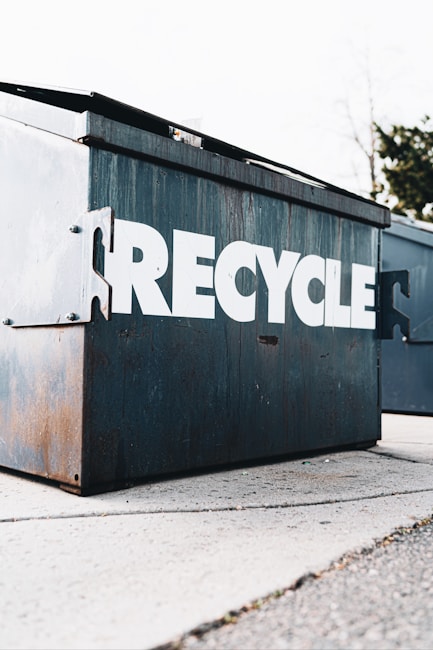 A large metal recycling container with the word 'RECYCLE' prominently displayed in white capital letters. The container shows signs of rust and wear, suggesting regular use. It is situated on a concrete surface, with trees visible in the background.
