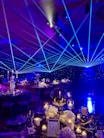 A formal event setup features tables adorned with elegant decorations including white flowers, glass candle holders, and a reflective centerpiece. Blue laser lights cross the room, creating a dynamic ceiling effect. The ambient lighting casts a starry atmosphere, while chairs are neatly arranged around each decorated dining area.