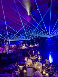 A formal event setup features tables adorned with elegant decorations including white flowers, glass candle holders, and a reflective centerpiece. Blue laser lights cross the room, creating a dynamic ceiling effect. The ambient lighting casts a starry atmosphere, while chairs are neatly arranged around each decorated dining area.