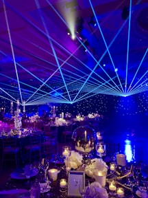 A formal event setup features tables adorned with elegant decorations including white flowers, glass candle holders, and a reflective centerpiece. Blue laser lights cross the room, creating a dynamic ceiling effect. The ambient lighting casts a starry atmosphere, while chairs are neatly arranged around each decorated dining area.