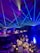 A formal event setup features tables adorned with elegant decorations including white flowers, glass candle holders, and a reflective centerpiece. Blue laser lights cross the room, creating a dynamic ceiling effect. The ambient lighting casts a starry atmosphere, while chairs are neatly arranged around each decorated dining area.