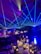 A formal event setup features tables adorned with elegant decorations including white flowers, glass candle holders, and a reflective centerpiece. Blue laser lights cross the room, creating a dynamic ceiling effect. The ambient lighting casts a starry atmosphere, while chairs are neatly arranged around each decorated dining area.