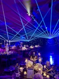 A formal event setup features tables adorned with elegant decorations including white flowers, glass candle holders, and a reflective centerpiece. Blue laser lights cross the room, creating a dynamic ceiling effect. The ambient lighting casts a starry atmosphere, while chairs are neatly arranged around each decorated dining area.