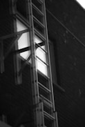 A metal ladder is positioned against the exterior wall of a building, rising up towards a window. The wall is constructed of dark brick, and the window reflects light, creating a contrast against the surrounding structure.