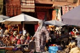 A bustling outdoor market scene with various colorful woven baskets and textiles displayed under large umbrellas. A person in a patterned outfit and headscarf walks among the stalls. The background includes vibrant rugs hanging on the walls and multiple people browsing the goods.