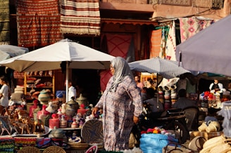 A bustling outdoor market scene with various colorful woven baskets and textiles displayed under large umbrellas. A person in a patterned outfit and headscarf walks among the stalls. The background includes vibrant rugs hanging on the walls and multiple people browsing the goods.