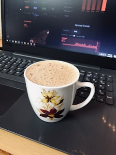 A white ceramic mug with floral designs sits on a laptop keyboard. The mug contains a frothy drink, possibly coffee or hot chocolate. The laptop screen displays graphic data and charts.