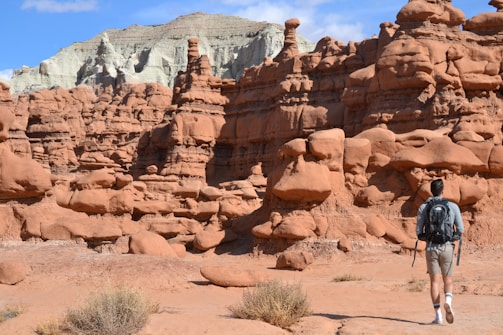 A person wearing a backpack and holding trekking poles is walking through a unique desert landscape filled with eroded sandstone formations and red rock hoodoos. The sky is clear with some clouds, and there are dry shrubs scattered on the ground.