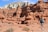 A person wearing a backpack and holding trekking poles is walking through a unique desert landscape filled with eroded sandstone formations and red rock hoodoos. The sky is clear with some clouds, and there are dry shrubs scattered on the ground.