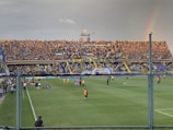 A large crowd fills a stadium stands, with vibrant yellow and blue banners and flags displayed prominently. A rainbow is visible in the sky to the right, adding an element of natural beauty to the scene. The field below has a few people dressed in sports attire, suggesting a sports event is taking place.