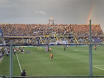 A large crowd fills a stadium stands, with vibrant yellow and blue banners and flags displayed prominently. A rainbow is visible in the sky to the right, adding an element of natural beauty to the scene. The field below has a few people dressed in sports attire, suggesting a sports event is taking place.