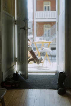 An open door leading to a sunlit porch with a small table and two chairs outside. A person is partially visible sitting on the porch, wearing boots and enjoying a beverage. Inside, a pair of shoes are placed on a doormat, suggesting a cozy indoor-outdoor atmosphere.