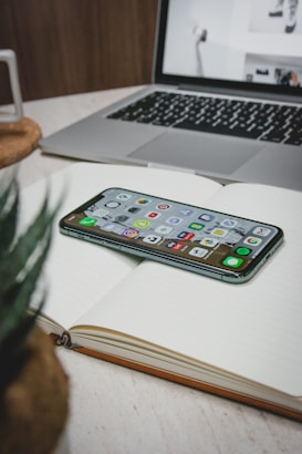 A smartphone displaying various app icons rests on an open notebook with lined pages. In the background, there is a laptop with a partially visible screen showing images. A plant in the foreground adds a touch of greenery to the scene.