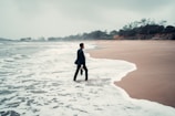 A person in business attire stands with one foot in the ocean surf on a deserted beach, holding a briefcase. The sky is overcast, and the surrounding scenery is lined with distant trees and greenery.