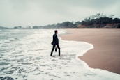 A person in business attire stands with one foot in the ocean surf on a deserted beach, holding a briefcase. The sky is overcast, and the surrounding scenery is lined with distant trees and greenery.