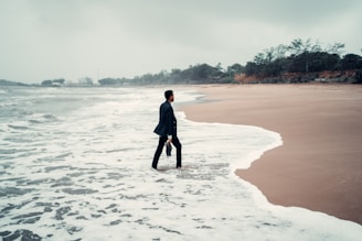 A person in business attire stands with one foot in the ocean surf on a deserted beach, holding a briefcase. The sky is overcast, and the surrounding scenery is lined with distant trees and greenery.
