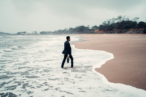 A person in business attire stands with one foot in the ocean surf on a deserted beach, holding a briefcase. The sky is overcast, and the surrounding scenery is lined with distant trees and greenery.