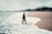A person in business attire stands with one foot in the ocean surf on a deserted beach, holding a briefcase. The sky is overcast, and the surrounding scenery is lined with distant trees and greenery.