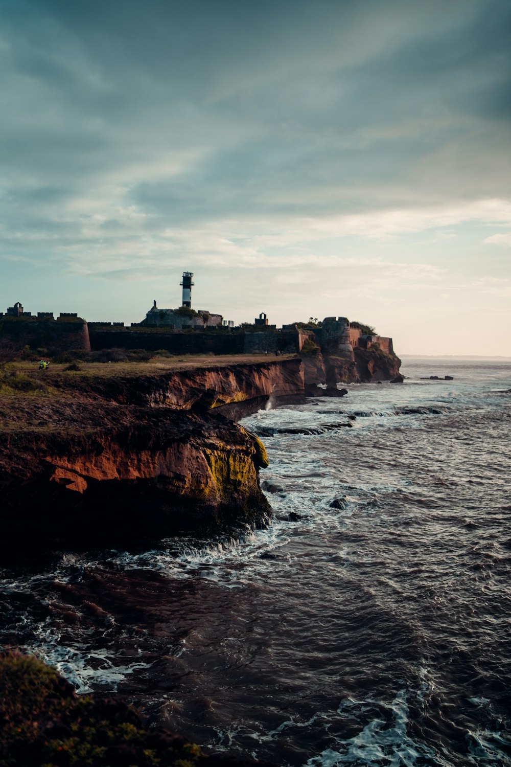 farol branco no penhasco marrom à beira-mar sob nuvens brancas durante o dia