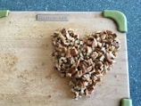 A heart shape made of chopped pecans is arranged on a wooden cutting board. The board has green handles on the corners, and it rests on a speckled countertop.