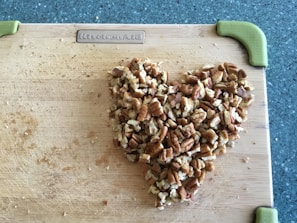 A heart shape made of chopped pecans is arranged on a wooden cutting board. The board has green handles on the corners, and it rests on a speckled countertop.
