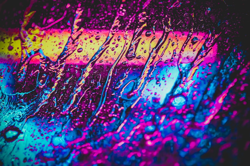 Vivid colors blend together in an abstract pattern resembling streaks of liquid or paint on glass. Bright and bold hues of pink, blue, and yellow create an eye-catching display that suggests motion and energy.