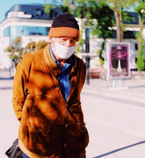 An elderly person is wearing a brown coat and a beanie, paired with a white face mask. The background reveals an urban setting with modern buildings and greenery.
