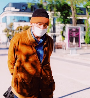 An elderly person is wearing a brown coat and a beanie, paired with a white face mask. The background reveals an urban setting with modern buildings and greenery.
