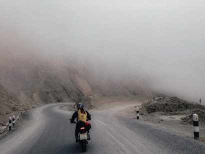 A motorcyclist is traveling on a winding road surrounded by a barren, rocky landscape. The sky is overcast, and fog obscures the mountains in the background, creating a mysterious and isolated atmosphere.