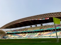 An outdoor stadium with a modern, curved roof structure and rows of seating in various colors. Flags from different countries are displayed in a line along the edge of the roof. A corner flag and portion of the grass field are visible in the foreground.