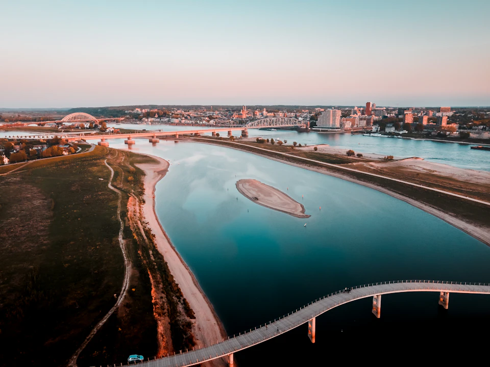 The image presents an aerial view of a river with a bridge spanning across it, connecting two parts of a city separated by the water body. The river is surrounded by buildings and trees on both sides, creating a picturesque urban landscape. The sky above has a light blue hue, suggesting either early morning or late afternoon hours.