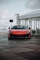 A sleek red sports car is parked on a wooden deck. The car has an aerodynamic design with a pronounced front bumper and sleek headlights. In the background, there is a white tent-like structure with glass walls and some lighting equipment. The sky appears cloudy.