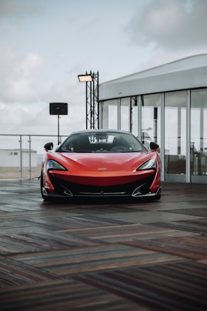A sleek red sports car is parked on a wooden deck. The car has an aerodynamic design with a pronounced front bumper and sleek headlights. In the background, there is a white tent-like structure with glass walls and some lighting equipment. The sky appears cloudy.