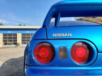A close-up of the rear end of a blue Nissan car, emphasizing its taillights and logo, set against a backdrop of an automotive garage with signs indicating 'import' and 'domestic'. The lighting is bright, highlighting the car's shiny blue paint.