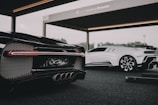 Two luxury sports cars are showcased in a modern, open garage. The foreground features a sleek black car with intricate tail light and exhaust details, while a white sports car stands in the background. The setting suggests a high-end automotive event or display.