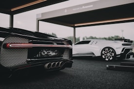 Two luxury sports cars are showcased in a modern, open garage. The foreground features a sleek black car with intricate tail light and exhaust details, while a white sports car stands in the background. The setting suggests a high-end automotive event or display.