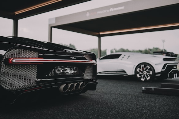 Two luxury sports cars are showcased in a modern, open garage. The foreground features a sleek black car with intricate tail light and exhaust details, while a white sports car stands in the background. The setting suggests a high-end automotive event or display.