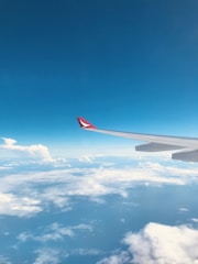 An airplane wing extends into the sky, with a clear view of fluffy white clouds below and a bright blue sky above. The wing tip shows a red marking or logo.